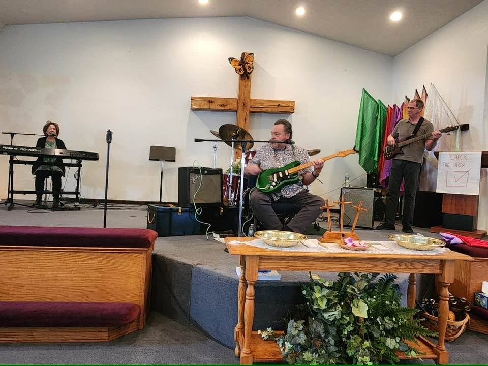 Worship team — keyboard, guitar and bass — leading praise on stage beneath the wooden cross